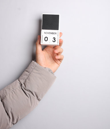 Man's hand in warm down jacket holds block calendar with date november 3 on gray backgroundの写真素材