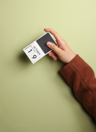Man's hand in coat holds block calendar with the date October 19 on green backgroundの写真素材
