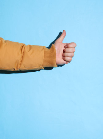 Male hand in a warm winter jacket showing a thumbs up on a blue background