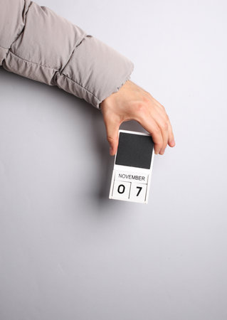 Man's hand in warm down jacket holds block calendar with date november 7 on gray background