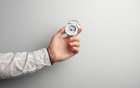 Man's hand in white shirt holds alarm clock on gray backgroundの写真素材