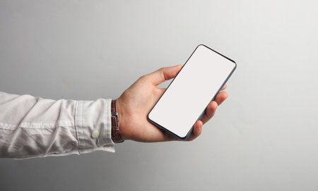 Man's hand in white shirt holds smartphone with white blank screen on gray background. Mockup, space for your informationの写真素材