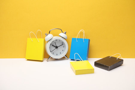 Miniature paper shopping bags and alarm clock on yellow gray background. Black Friday, world shopping day, holiday sale conceptの写真素材