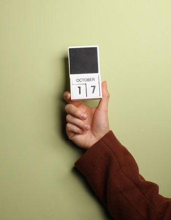 Man's hand in coat holds block calendar with the date October 17 on green backgroundの写真素材