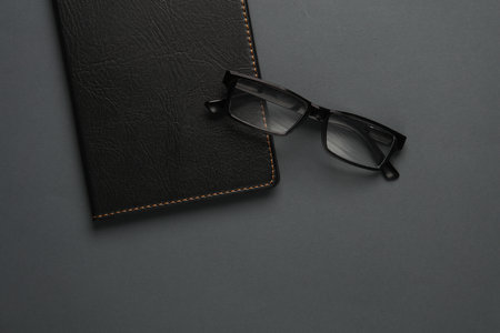 Notebook with black leather cover and eyeglasses on dark gray background. Business concept. Top...