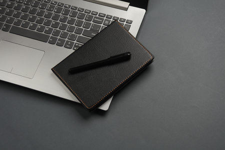 Laptop and Notebook with black leather cover, pen on dark gray background