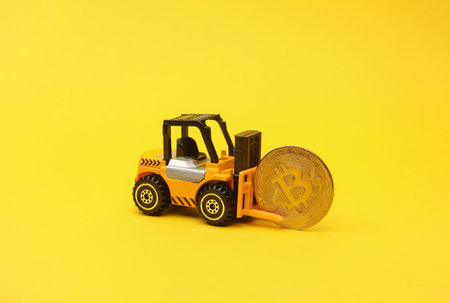 Miniature toy forklift and bitcoin on a yellow background.