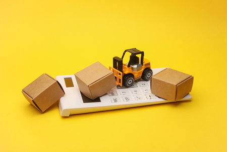 Miniature toy forklift and calculator with boxes on a yellow background. Logistics, transportation,...