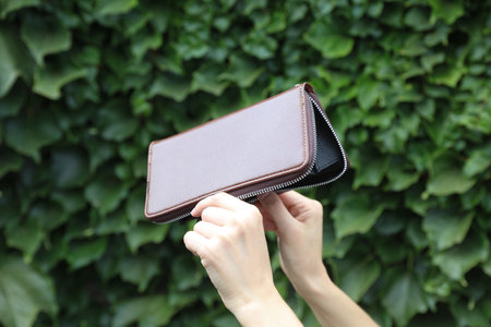 Brown leather purse in woman's hands against the green leaves wall background