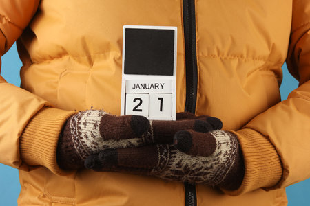 Man in yellow winter down jacket and gloves holding block calendar with date January 21 on blue backgroundの写真素材