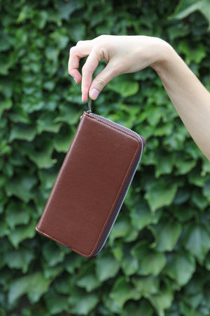 Brown leather purse in woman's hand against the green leaves wall background