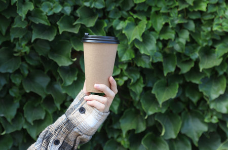 Hand in shirt holds take out coffee cup on green leaves wall background