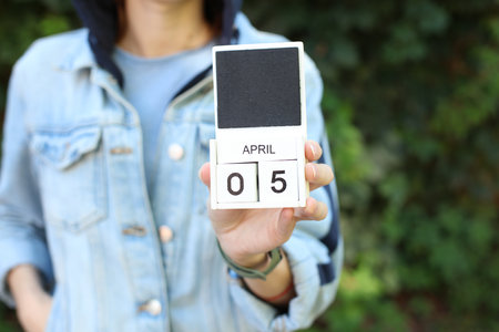 Woman in denim jacket holds white block wooden calendar with date April 5 outdoors