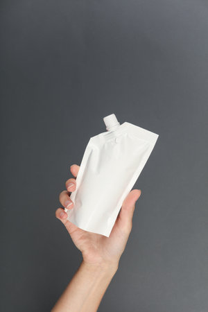 Hand holds white doy pack with dispenser on a dark gray background. Template for design, mockup