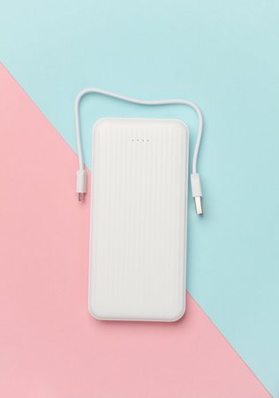 White power bank with cable on pink blue background. External battery for charging smartphone and...