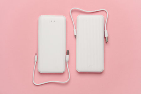 White power banks with cable on pink background. External batteries for charging smartphone and...