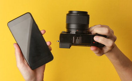 Hands holding a modern mirrorless camera and smartphone on yellow backgroundの写真素材