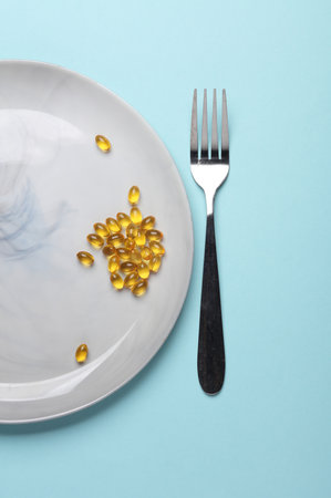 Fish oil capsules in plate with fork on blue background. Diet, healthy eatingの写真素材