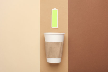 Cardboard coffee cup with a fully charged battery on a beige background. Charge of energy and strength for the whole dayの写真素材