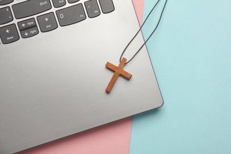 Wooden Christian cross with laptop on pink blue background. Online Bible study. Flat lay