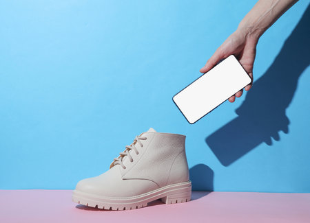 Women's white leather winter boot and hand holding smartphone on pink blue background with shadow. Creative layout. Minimalism fashion still life. Mockupの写真素材