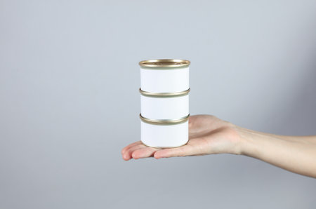 Tin cans of canned food with a white label, mockup for your design in a female hand on a gray studio...