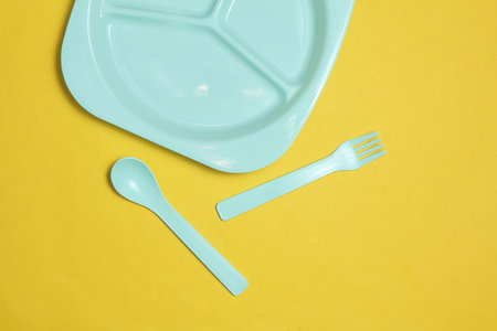 Separate plastic plate with spoon and fork for feeding baby on yellow background. Top view