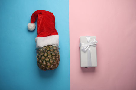 Pineapple in santa hat and gift box on pink blue background. Merry Christmas conceptの写真素材