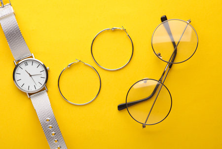 Stylish wristwatch with eyeglasses and earrings on yellow background. Women's accessories. Top viewの写真素材