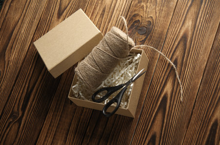 DIY, Product packaging, presentation. Kraft cardboard box with filler, scissors and thread on wooden...