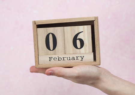 Woman's hand holds wooden block calendar with the date February 06 on pink concrete background