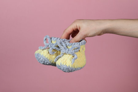 Woman's hand holds baby knitted shoes for newborn on a pink backgroundの写真素材