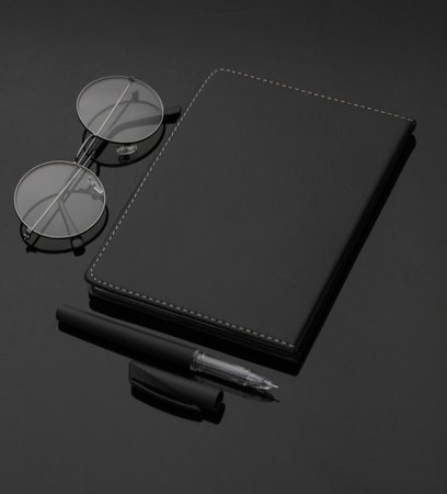 Black leather notebook with eyeglasses and pen on a black backgroundの写真素材