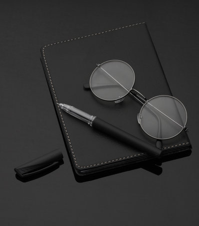 Black leather notebook with eyeglasses and pen on a black backgroundの写真素材