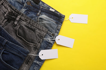 Assortment of many different fashionable jeans pants with blank paper tags. Shopping, sale concept. Yellow backgroundの写真素材