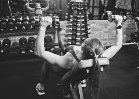 Young woman doing exercise with dumbbells for the muscles of chest and shoulders on inclined bench in the gym. Back view. Black and whiteの写真素材
