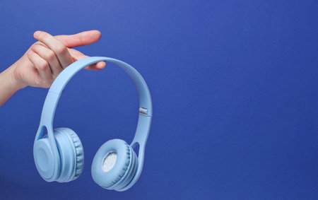 Modern blue wireless headphones on finger of female hand on blue background. Copy spaceの写真素材