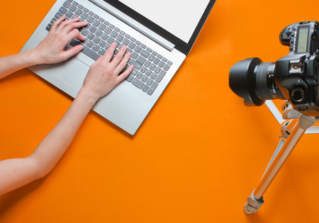 Woman blogger typing on a laptop, blogging with a camera with a tripod on orange background....