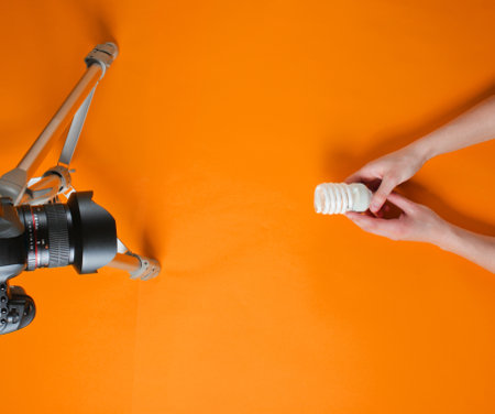 Women's hand review eco spiral light bulb with camera on tripod on orange background. Top view. Minimalismの写真素材