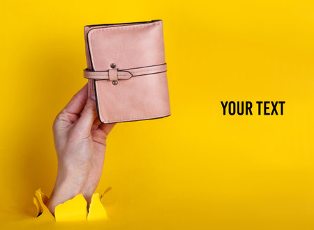 Female hand holding pink wallet through torn yellow paper. Minimalistic creative fashion concept. Copy spaceの写真素材
