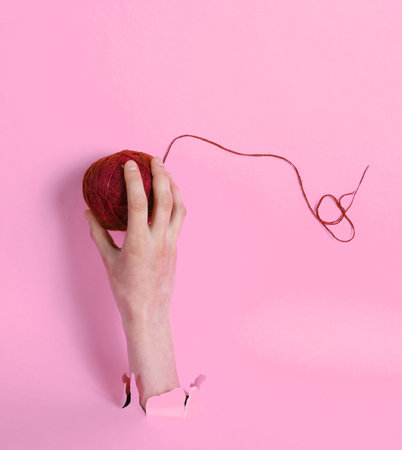 Female hand holding skein of thread through torn pink paper. Minimalistic creative concept