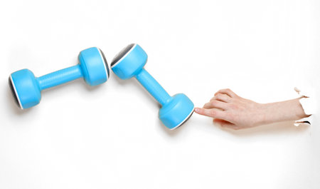 Female hand takes blue plastic dumbbell through torn white paper. Minimalistic sport conceptの写真素材