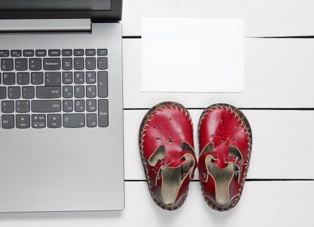 Laptop with white piece of paper for copy space and kids sandals on wooden background, top view.の写真素材