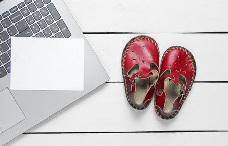 Laptop with white piece of paper for copy space and kids sandals on wooden background, top view.