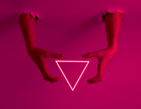 Female hands through the torn holes with neon triangle
. Pink light. Copy space. Creative artの写真素材
