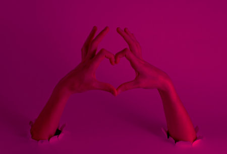 Female hands show a heart symbol through the torn holes with pink neon light. Creative artの写真素材