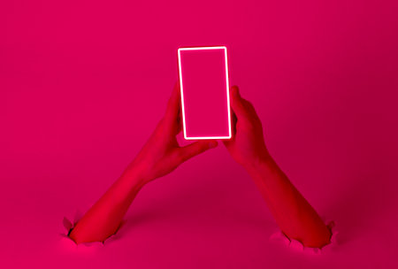 Female hands holding smartphone with neon glow frame through the torn holes. Pink light. creative artの写真素材