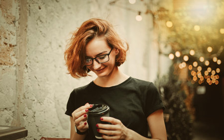 Cheerful hipster girl in glasses with short haircuts holding coffee cup against the background of...