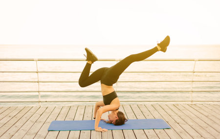 Young woman doing yoga exercises standing on her head on a mat at the beach