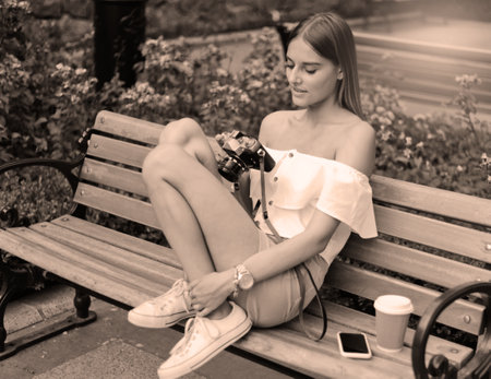 Young beautiful woman in summer clothes holding a retro camera while sitting on bench in the parkの写真素材